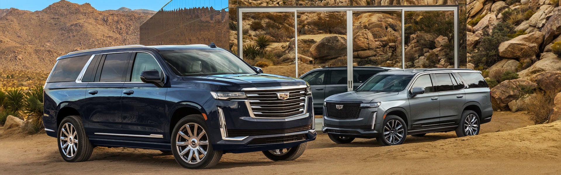 Two Cadillac Escalades parked with rocky mountains and a glassy building in the background
